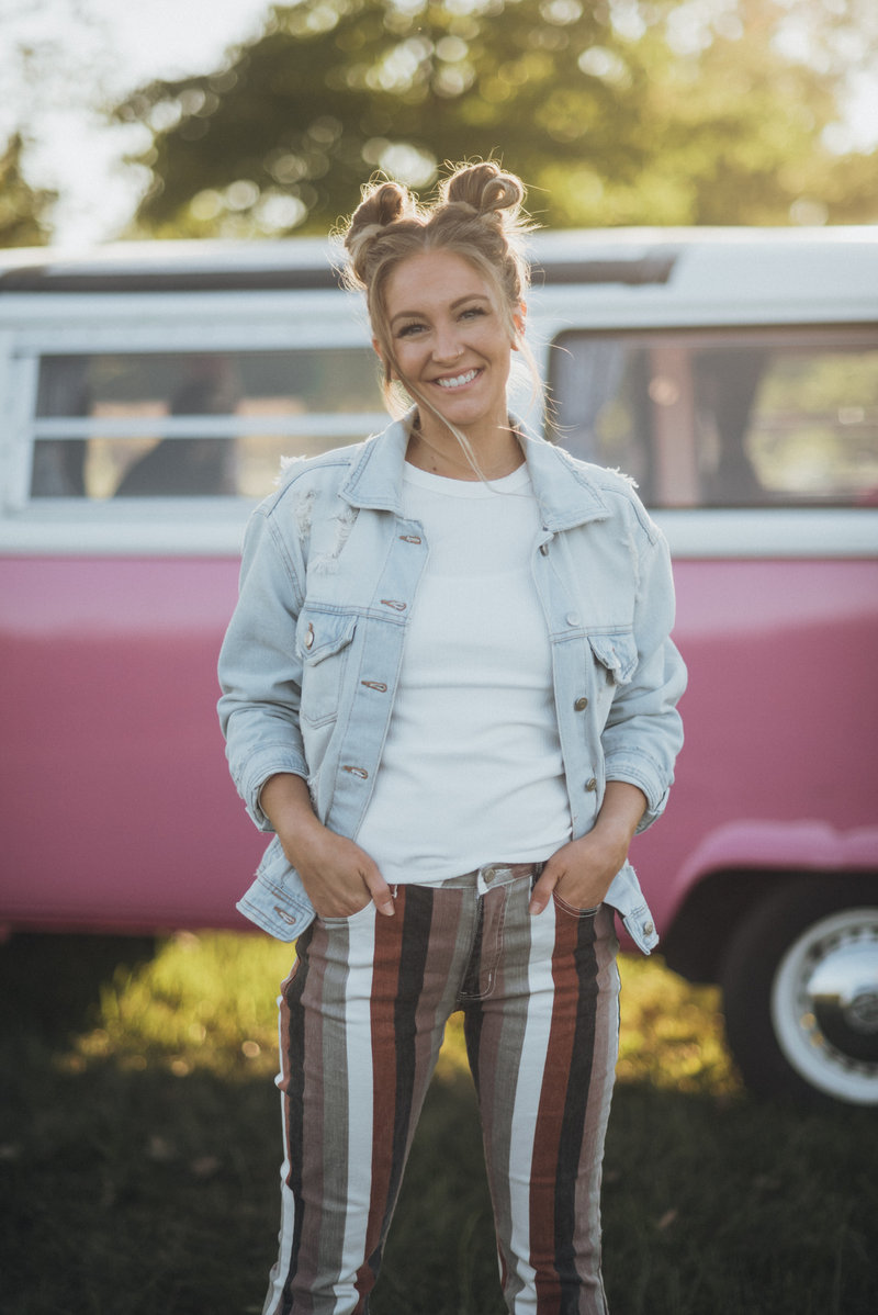 Liz smiling in a denim jacket in front of a pink vintage VW bus at golden hour