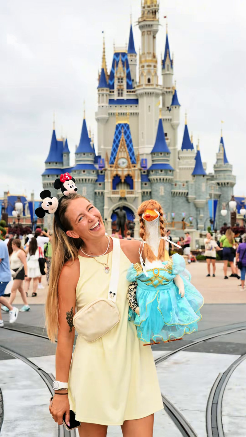 Liz laughing in Mickey and Minnie ears, holding Lucy Goosey in her ballgown in front of Cinderella Castle