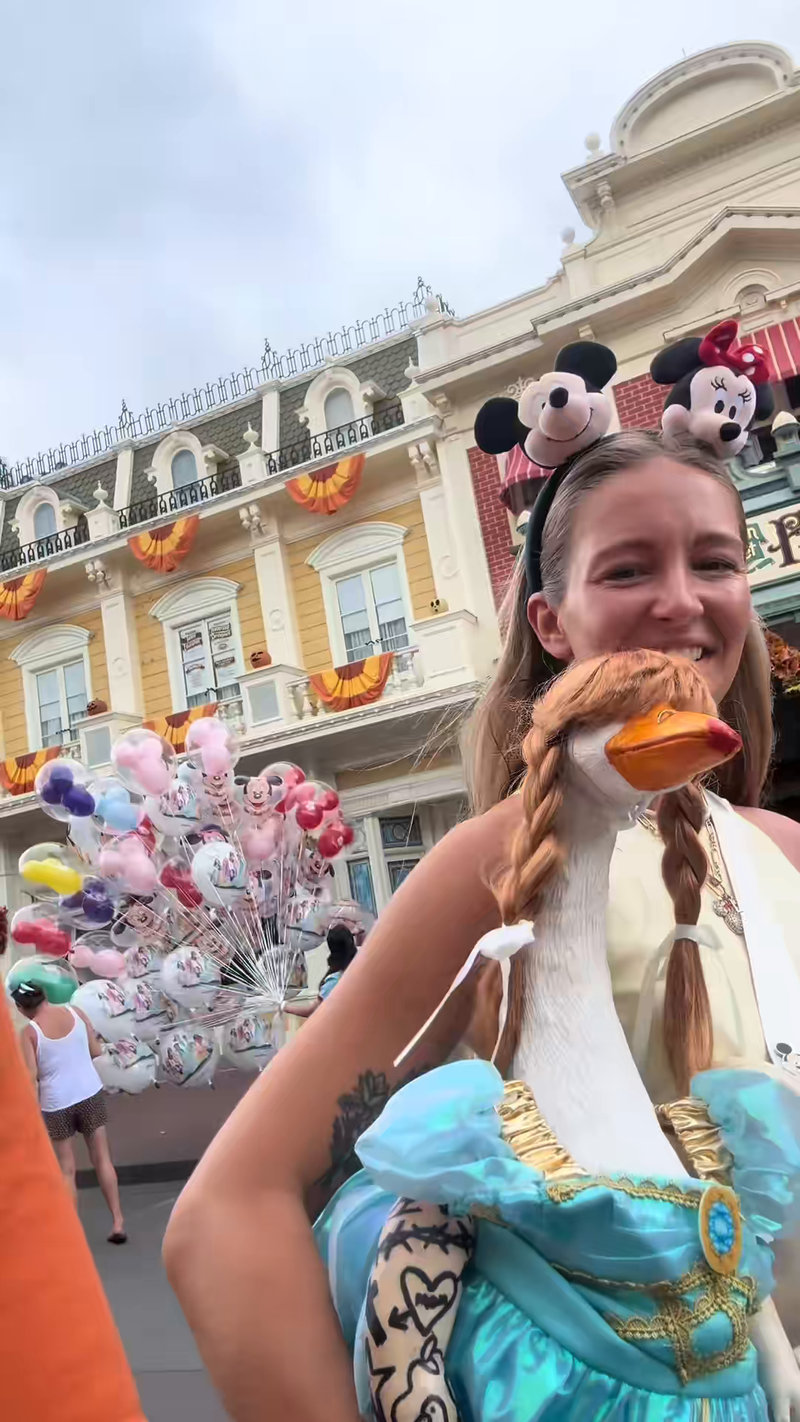 Liz smiling on Main Street at Magic Kingdom with Lucy Goosey on her shoulder, a balloon seller behind them