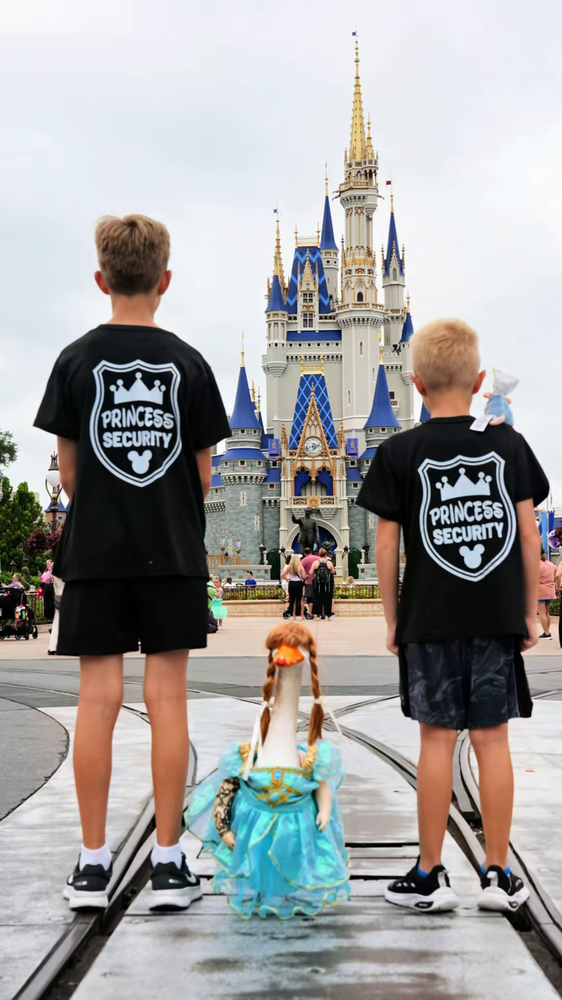 Two boys seen from behind in black Princess Security shirts standing guard either side of Lucy Goosey in front of Cinderella Castle